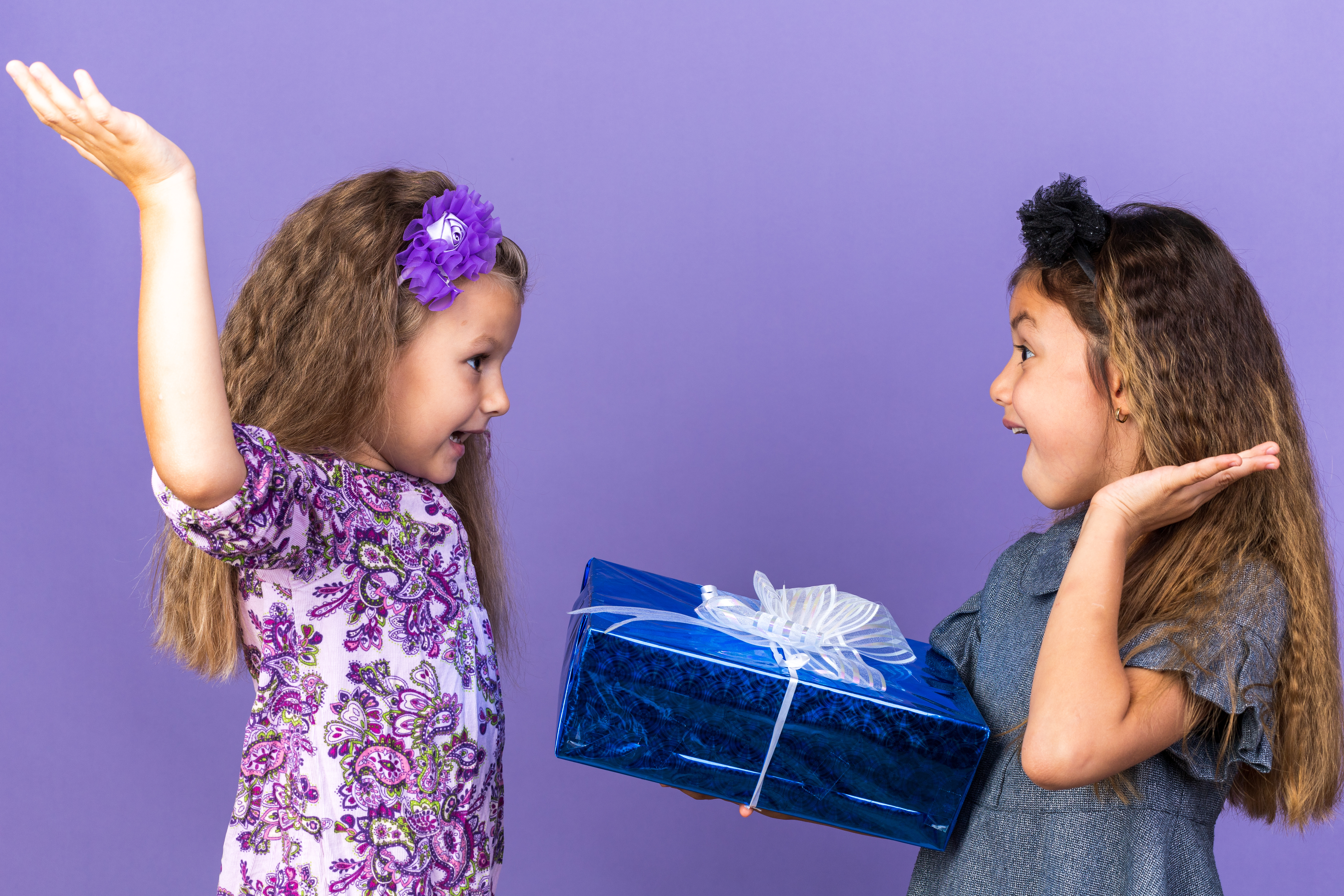 surprised-little-blonde-girl-standing-with-raised-hands-looking-her-friend-holding-gift-box-isolated-purple-wall-with-copy-space-1